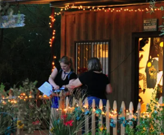 Two people interacting outside decorated wooden building with string lights and flower-adorned fence.