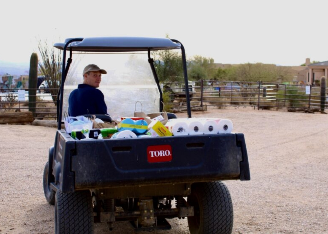 Person driving a utility vehicle loaded with supplies on gravel in desert setting.