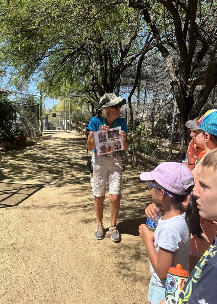 Tour guide showing photos to a group of children at a wildlife facility on a sunny day.